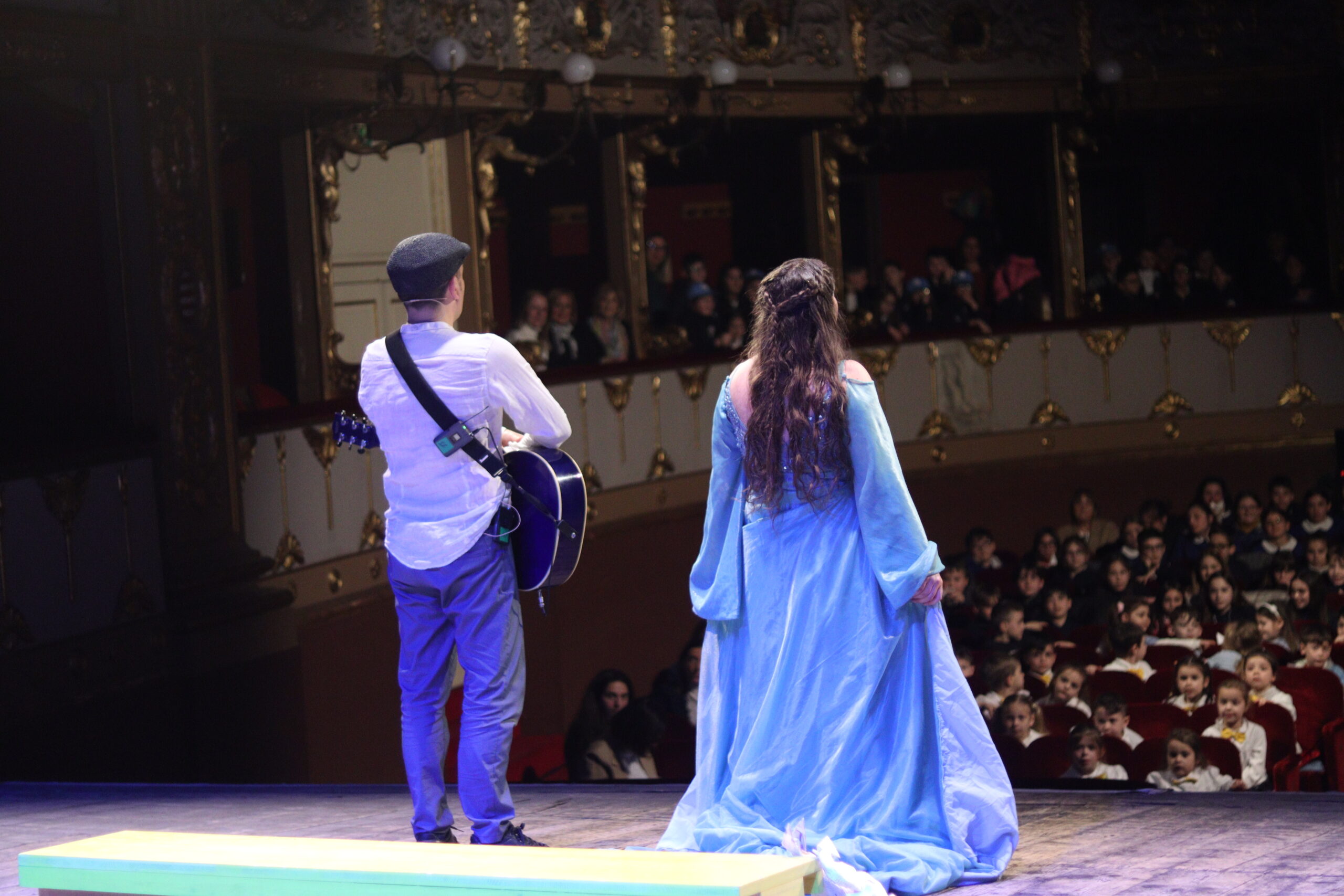 Attori di Room to Play in scena con chitarra e abito azzurro davanti al pubblico di un teatro